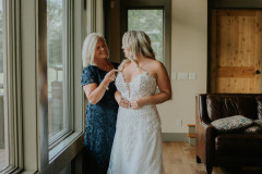bride being helped by mom put on dress in front of big windows.