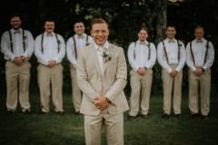 groom in tan suit standing in front of 6 groomsmen without jackets on.
