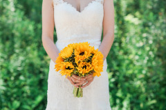bride in white wedding dress standing in front of greenery holding a bouquet made of large yellow sunflowers.