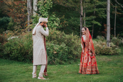 man in tradiitonal Indian garb holding hands over eyes facing toward woman in traditional Indian garb (red and gold decorations) - both standing in grassy area by woods.