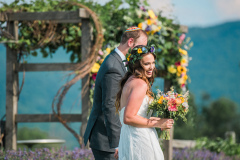 Man and woman in wedding attire holding hands in front of floral arrangement hanging on wooden fixture outdoors at Mountain Top Inn & Resort in Chittenden, VT