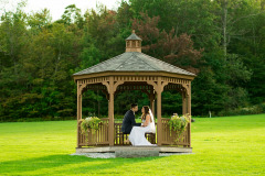 a wooden gazebo in a green meadow with green trees in the background.  a bride and groom sitting inside the gazebo.