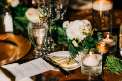 wooden table set with candles floating in water filled hurricanes, green and white flowers, a table card and gold chargers.