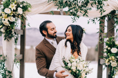 bride in white gown with veil, groom with beard in brown suit standing by arbor adorned with white and green flowers and white drapery.