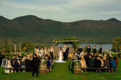 Group of people at outdoor wedding in various attire sitting on wooden benches in front of floral archway with bridal party and groom party in front overlooking lake and mountain at Mountain Top Inn & Resort in Chittenden, VT