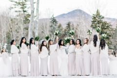 a row of bridesmaids with a bride in the center, with their backs to the camera but turning their heads to face it, holding up their white flower bouquets with a snowy mountain and woods backdrop.