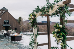 a wooden arbor adorned with white and green flowers and white drapery.  a brown wooden barn in the background and snowy grounds with mountains beyond.