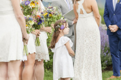 Partial shot of bridesmaids in outdoor wedding ceremony wearing white summer dresses and holding multi-colored bouquets.  A young flower girl with a flower crown in her long brown hair stands behind the bride.