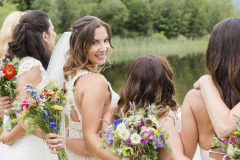 smiling bride in white with veil holding flowers behind bridesmaids to her side and looking back over should smiling.