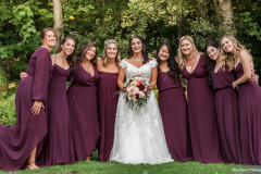8 bridesmaids wearing burgundy dresses flanking a brunette bride wearing a traditional wedding dress holding a burgundy and pink bouquet.  green trees in background.