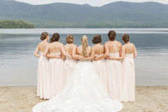 7 woman wedding  party with bride in center in white gown flanked by 6 women in pale pink formal dresses standing on beach in front of lake and mountains.