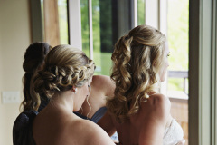 Blonde bride with ringlets in hair being helped by blond bridesmaid with dress.