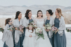 snowcapped mountains in background.  Bride in traditional white dress standing with flower girl in white dress and flanked by 5 bridesmaids in grey dresses.