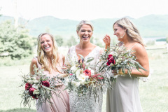 Blonde bride in lacy white dress standing with 2  blonde bridesmaids in front of green mountains.  Both women are holding oversized bouquets with red, white and green colors.