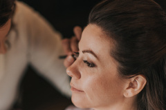 brunette with hair pulled back having her eye makeup done by a woman wearing a white shirt.
