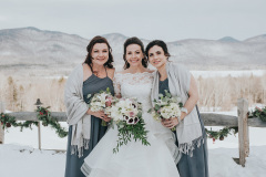 bride in traditional white gown holding burgundy and white bouquet, flanked by 2 bridesmaids in grey dresses with white shawls - standing in snow with fence and mountains in background.