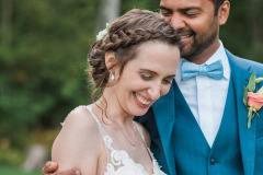brunette with hair up in braids wearing a strappy white dress receiving a kiss from a man in a blue suit.