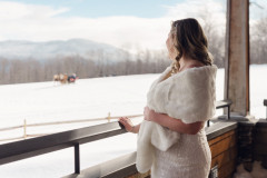 bride in white dress and white shawl on a deck of a house, overlooking a white field where a blurry sleigh ride is taking place. mountains in background.