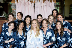 bride sitting with 11 bridesmaids all in blue floral robes in front of pink and white dresses in background smiling