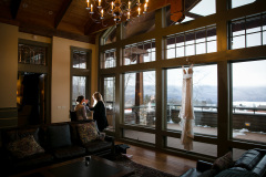 Room with large candelabra chandelier and large windows with view out to snowy mountains.  Wedding dress hanging in front of window and 2 women working on makeup in the background.
