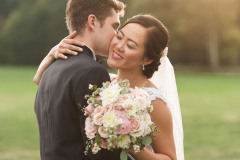 bride and groom hugging with greenery in background. groom wearing a suite, bride wearing a white dress and veil with pink and white bouquet.