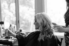 black and white photo of blonde  woman  in black cape from side profile  looking into a handheld mirror while another woman works on her hair.