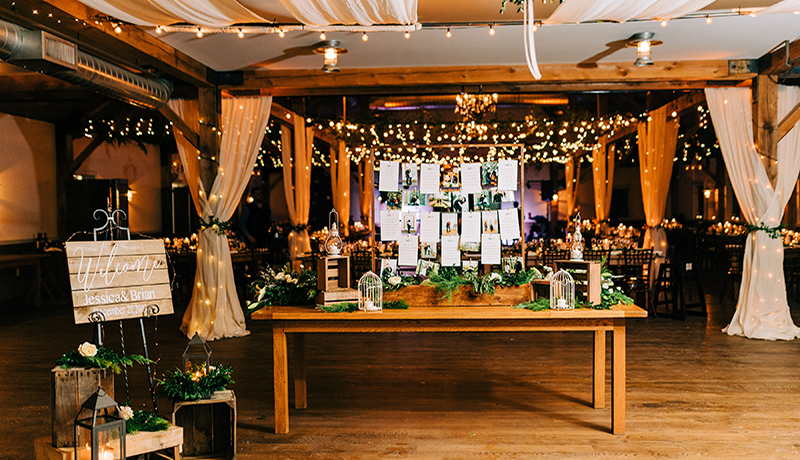 farmtable welcome table with name cards, greenery, lanterns and gifts. room decorated with greens and cream drapery.
