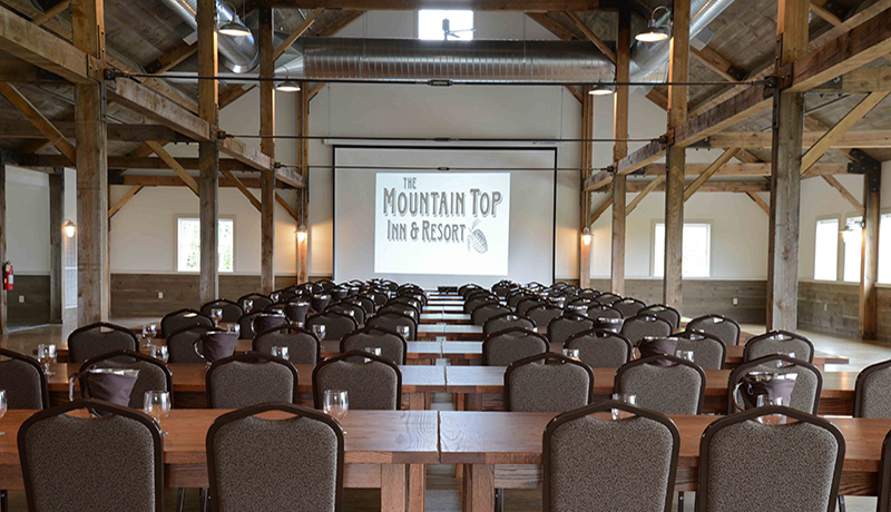 room with white walls and exposed wood beams and metal ductwork with rows of grey meeting chairs and wooden tables. projector screen at end of room.