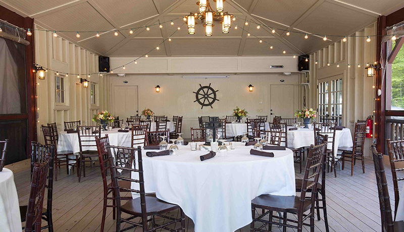 room filled with round tables with white table clothes and set for dinner with wooden chairs. chandelier in middle of ceiling, white walls and windows on left and right.