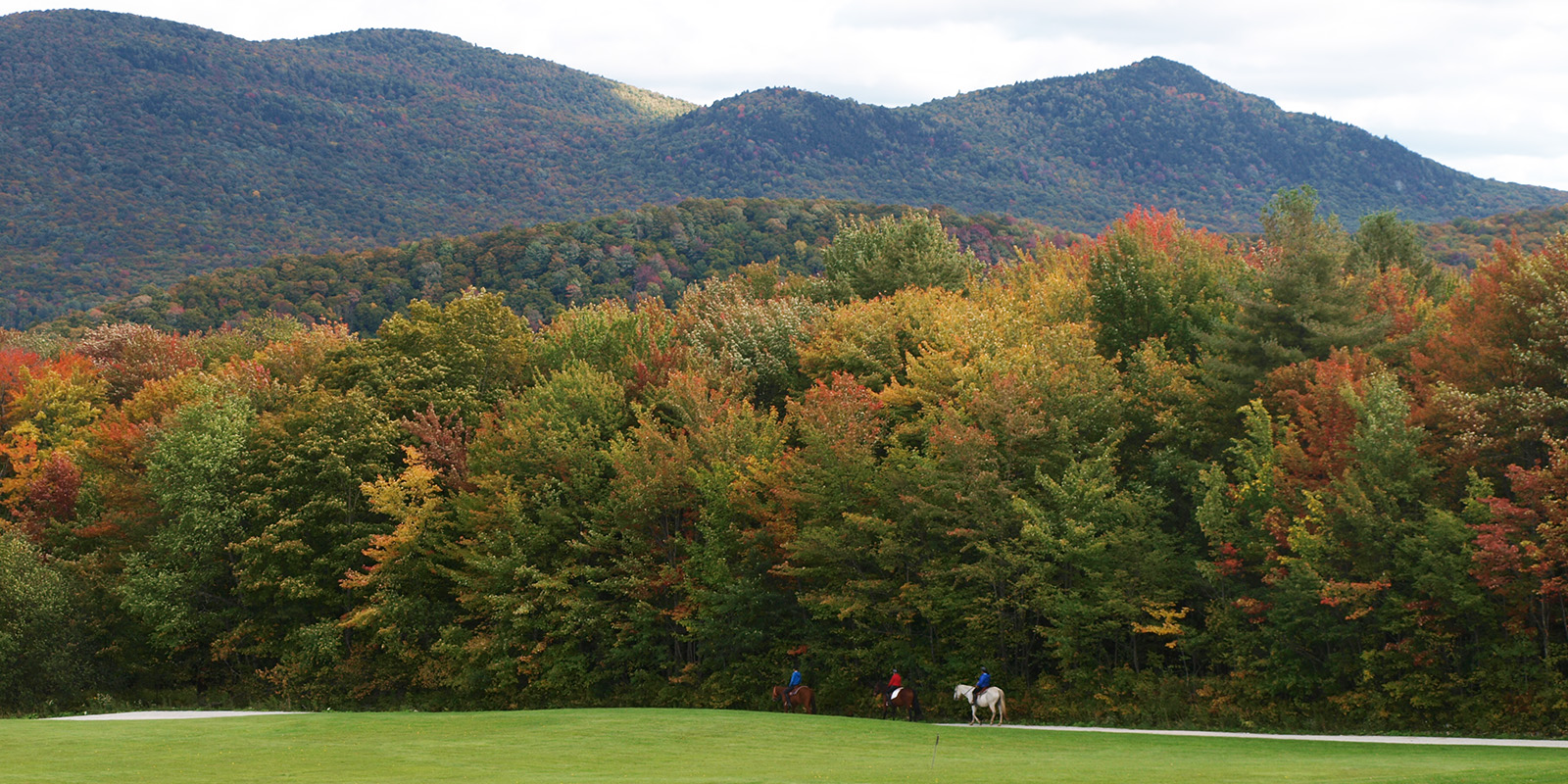 3 riders on horses along brightly colored fall woods with grass in the foreground and tree covered mountains in the background