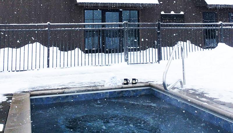 outdoor hot tub surrounded by snow in front of a building 2 cups of cocoa at the water's edge