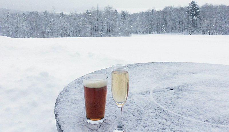 Pint of amber beer and champagne flute on snow covered table with snowy field and trees in background