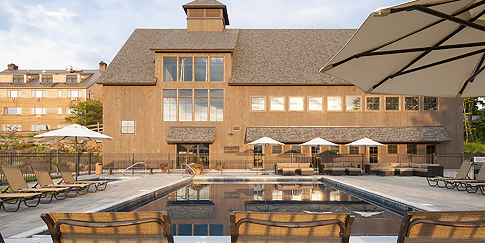 wooden exteriors of 3 story barn with windows and lodge in background, pool with tan lounge chairs and cream umbrellas in foreground