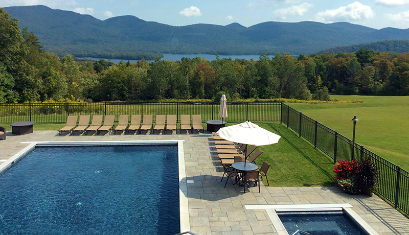 view from upper window of barn building looking out toward rectangular inground pool and hot tubs with slate patios and tan lounge chairs and white standing umbrellas surrounded by metal fence and grass with summer trees, lake and mountains in background. blue sky dotted with clouds.