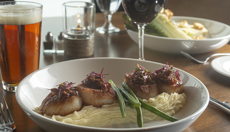 dish of scallops, mashed potatoes and string beans on a wood table with red wine
