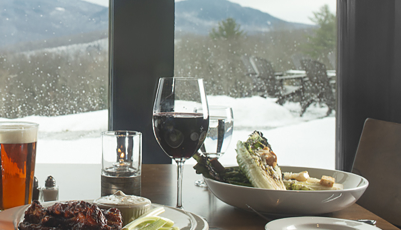 glass of red wine on a table with a ceasar salad and other food and snowy view out window