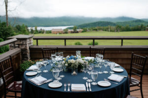 green mountains with lake in background, with lodge in midground, with blue table cloth and table setting in foreground with flora.