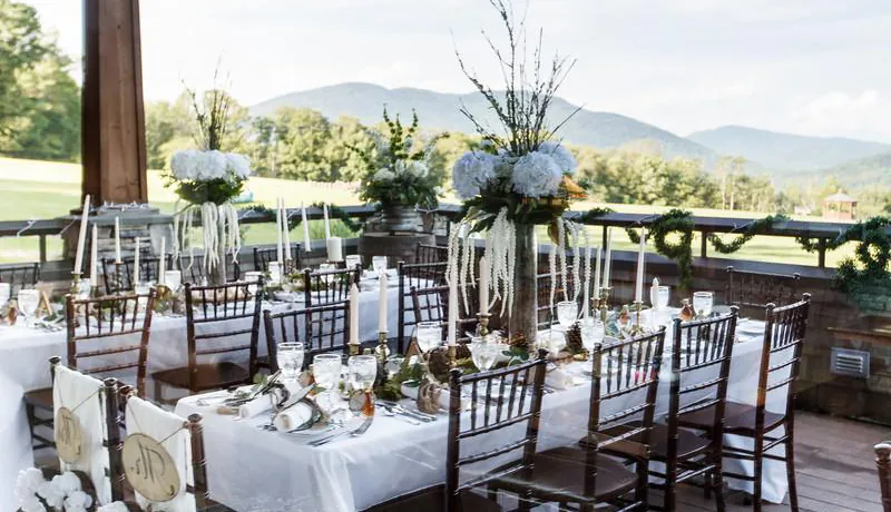 Photograph of Mountain Top Resort Guest Home, the Jewel, prepared for a wedding reception. Featuring two large farmhouse tables with chivari chairs, and white linens on tables and florals and greenery. 