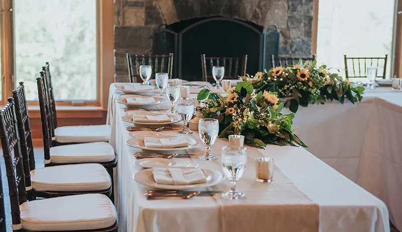 room with rectangular tables with white table clothes, white china and glasses, white cushions on wooden chairs, yellow flowers with greens as table decoration and fireplace and window in background