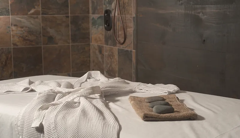 massage service table with white linens and white waffle stitch robe draped over it, brown tile shower wall in background and hot massage stones on a tan towel beside the robe.