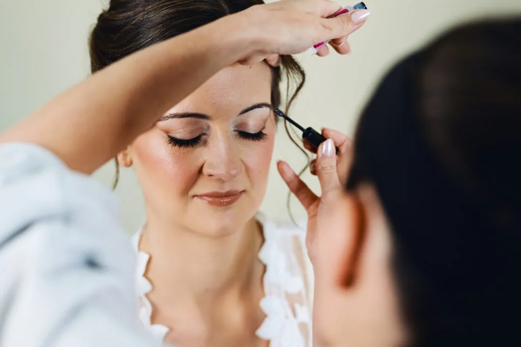 Bride in foreground getting makeup done by a makeup artist. You can see the makeup artists hands as she applies liner to the bride's eyebrows.